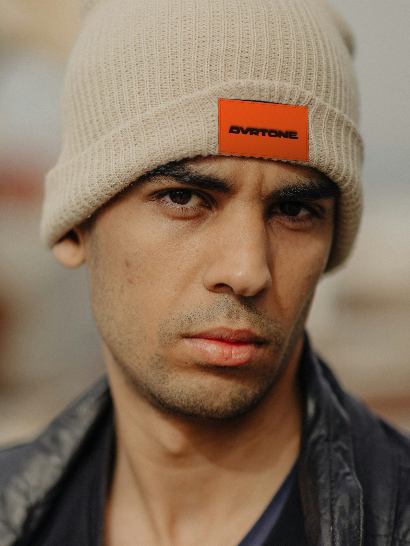 A man in an oat ribbed beanie with an orange woven OVRTONE patch on the cuff