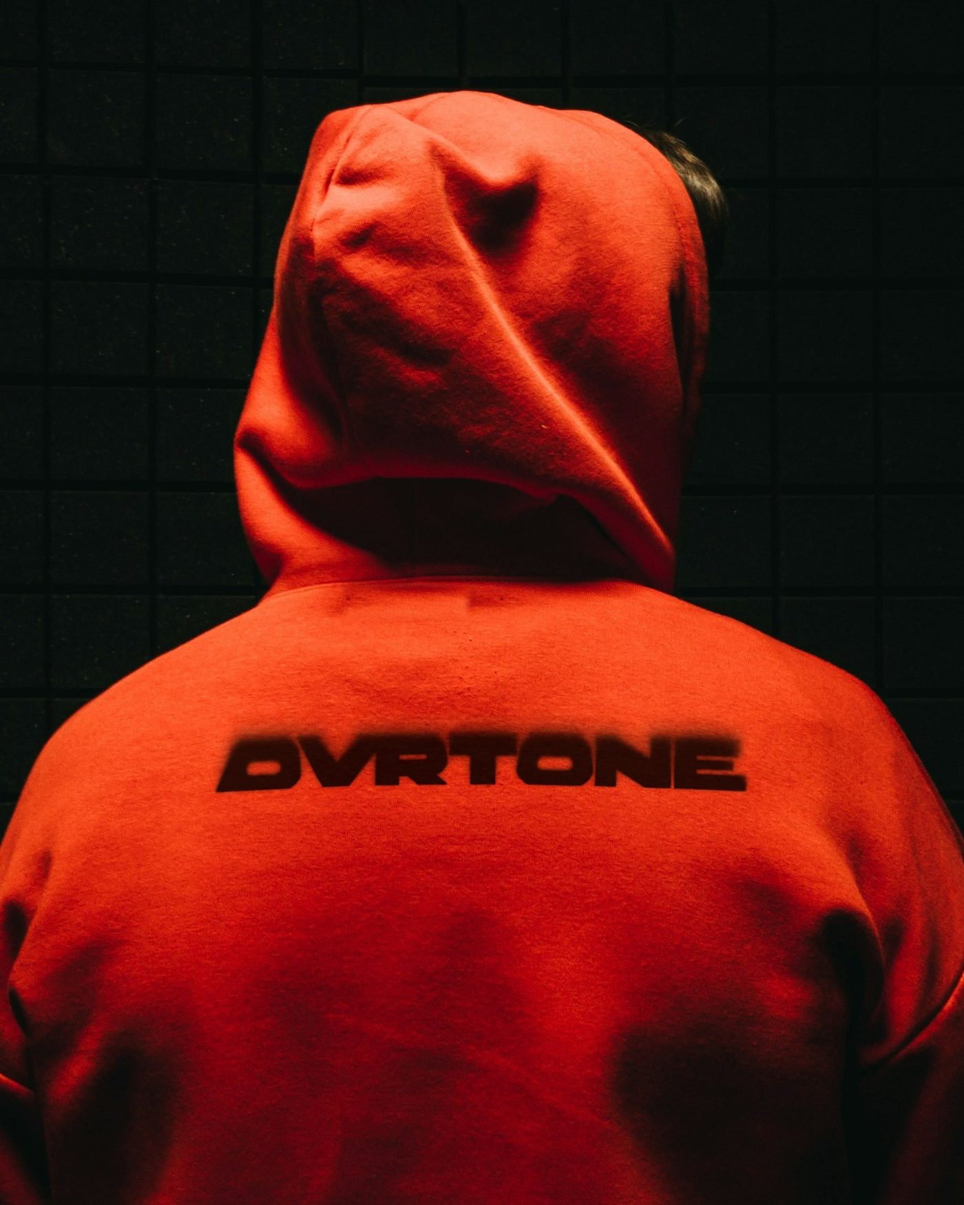 From behind, someone in an orange hoodie with the hood up, the blurred OVRTONE wordmark in black across the back