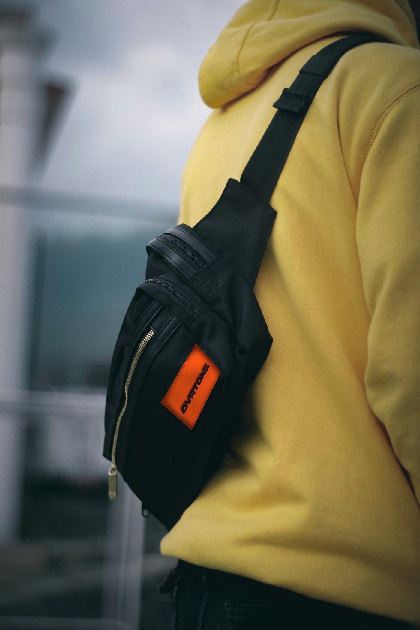 A black sling bag over a yellow hoodie, with an orange woven OVRTONE patch