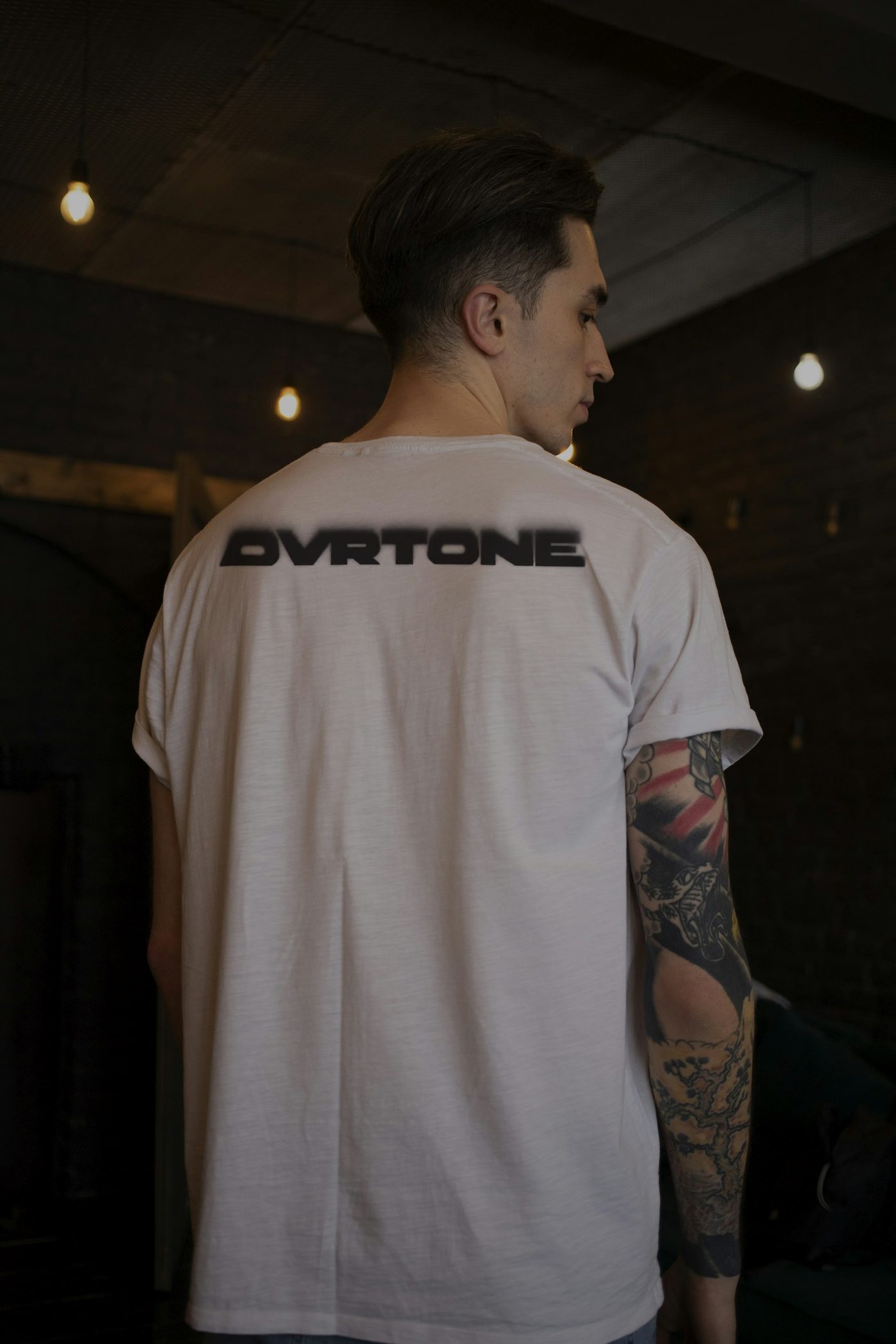 From behind, a man in a bone-white tee with the blurred OVRTONE wordmark in black across his upper back